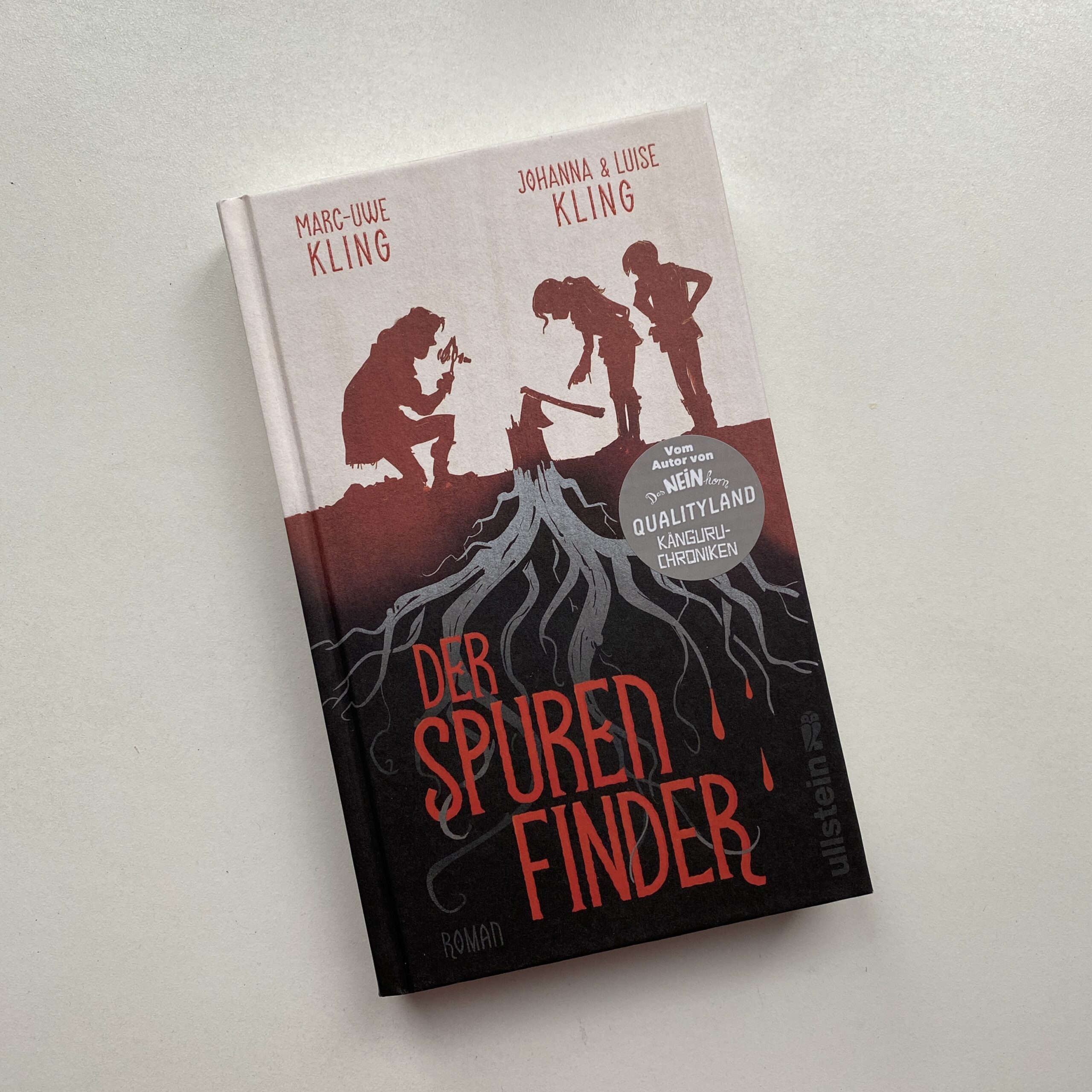 Foto des Buches "Spurenfinder" auf weißem Grund. Das Cover zeigt rote Scherenschnitt-Figuren die sich über eine Axt beugen und das Wurzelgeflecht des Baumstumpfes, in dem die Axt steckt.