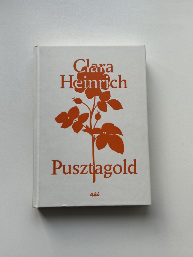 Buch auf weißem Grund, das Cover zeigt eine orangefarbene Blume auf weiß. Dazu der Schriftzug "Clara Heinrich" und "Pustzagold"